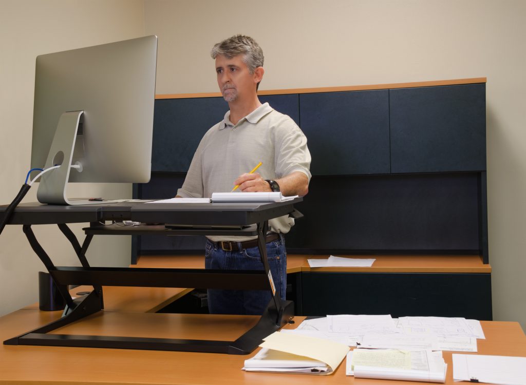 A man is working at a standup desk in an office where he works because standing is healthier than sitting all day. Live healthy, don’t sit all day.